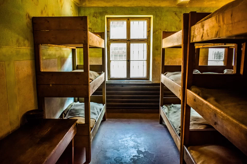 Zdjęcie przedstawia pomieszczenie z dwoma oknami, w którym stoją trzy trzypiętrowe prycze oraz drewniany stół. W oknach są kraty. Ściany są zielono‑żółte, a podłoga jest betonowa.
