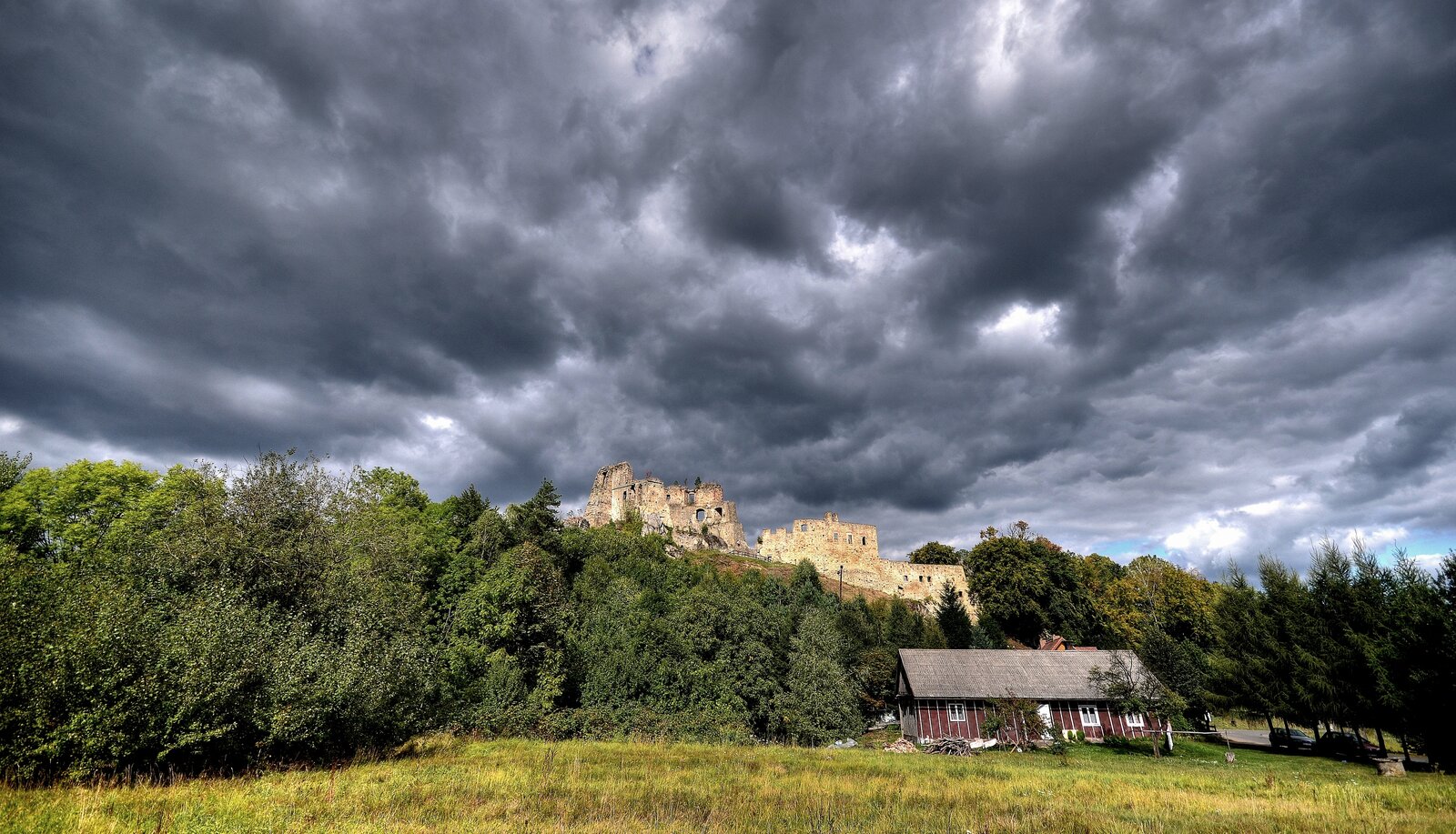 Zdjęcie przedstawia ruiny zamku i baszty stojące na wzgórzu, nad którymi kłębią się ciemne burzowe chmury. Wokół wzgórza rosną drzewa, a u jego stóp stoi drewniana, brązowa chata, przy której przebiega droga. Na niej stoją samochody.