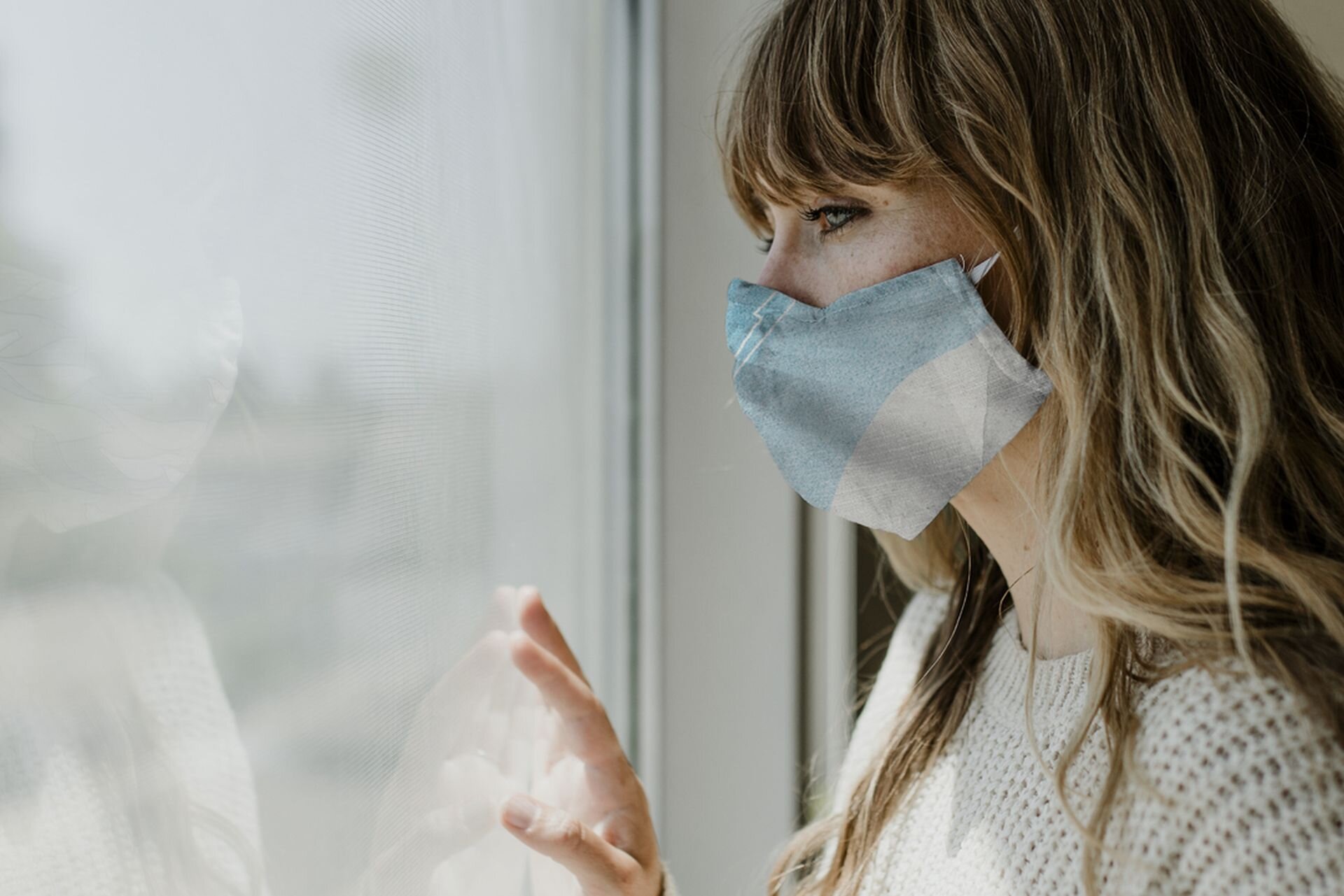 Zdjęcie przedstawia młoda kobietę z maseczką na twarzy, spoglądającą przez okno mieszkania.