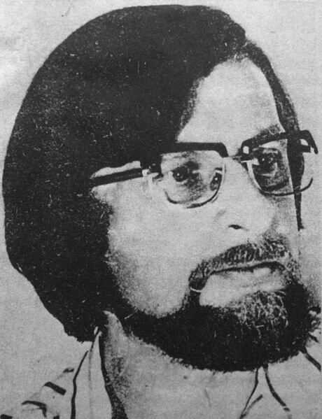 Zdjęcie przedstawia portret mężczyzny w ciemnych dłuższych włosach, w okularach i z zarostem. Ma poważny wyraz twarzy.