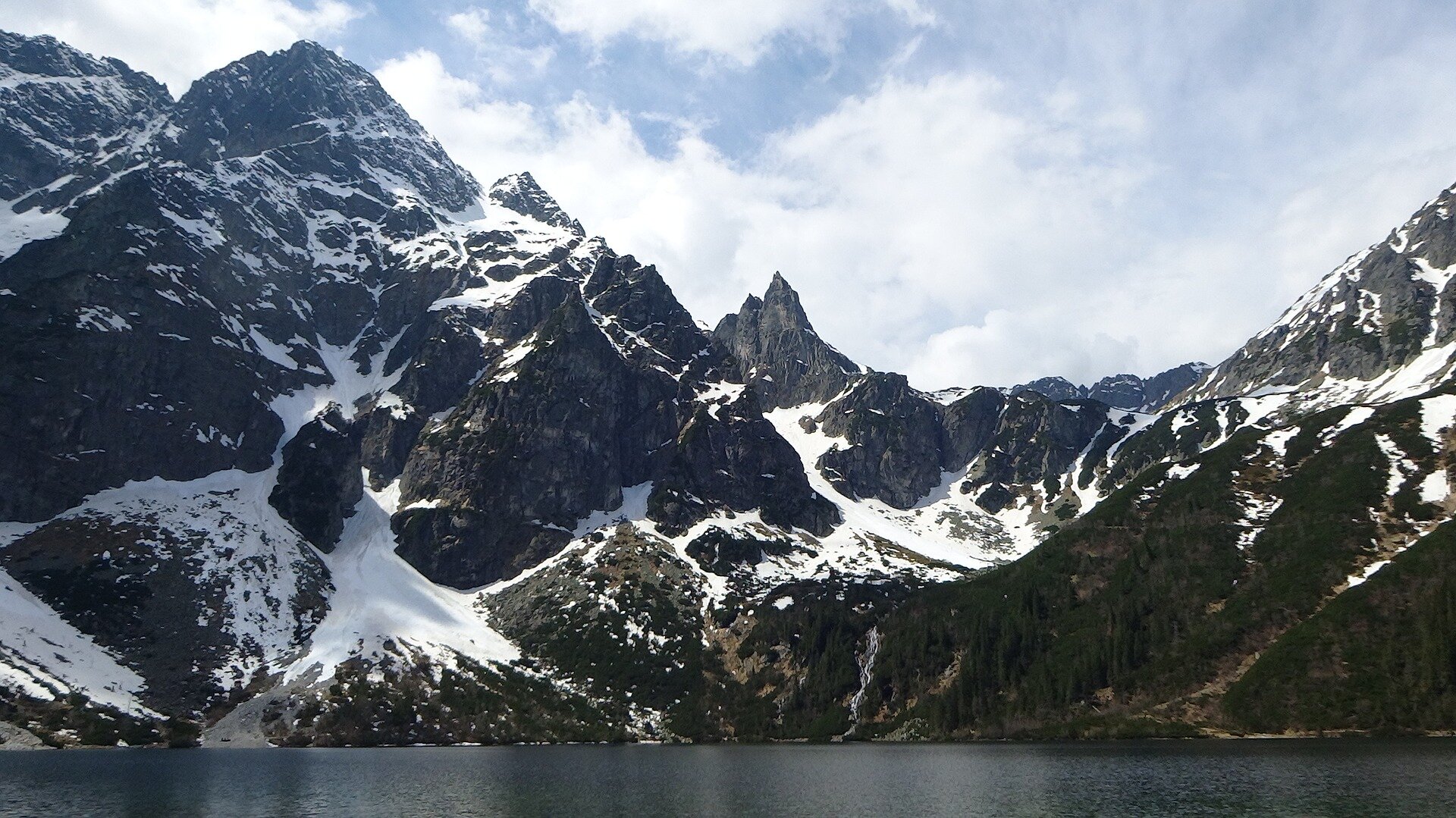 Kliknij, aby powiększyć. Zorientowana poziomo fotografia przedstawiająca krajobraz górski. Na pierwszym planie znajduje się jezioro o ciemnogranatowej, prawie czarnej, barwie. Za nim wysokie ukazane zostały wysokie góry, które są częściowo pokryte śniegiem. Nad nimi widnieje zachmurzone niebo.