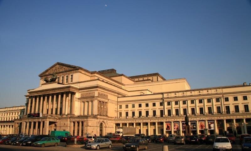 Zdjęcie przedstawia monumentalny budynek. Centralna jego część jest wysunięta do przodu i składa się z trzech pięter. Dwa pierwsze są wsparte kolumnami. Na dachu pierwszego piętra rzeźba. Z obu stron budynku niższe, podłużne skrzydła. Dach na parterze wsparty kolumnami. Przed budynkiem zaparkowane różnokolorowe samochody osobowe. Niebo błękitne, bezchmurne. Zdjęcie zostało wykonane w świetle dziennym.
