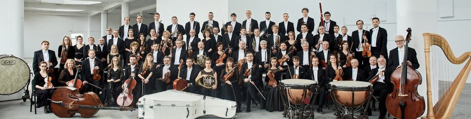 Ilustracja przedstawia zdjęcie Sinfonii Varsovii. Na kolorowej fotografii widoczna jest orkiestra, ustawiona w czterech rzędach. Na zdjęciu znajduje się blisko siedemdziesiąt osób. Muzycy ukazani są wraz z instrumentami. Po bokach widzimy instrumenty, których muzycy, z racji rozmiarów nie mogli trzymać przy sobie: bęben wielki, kontrabas, kotły, harfa.