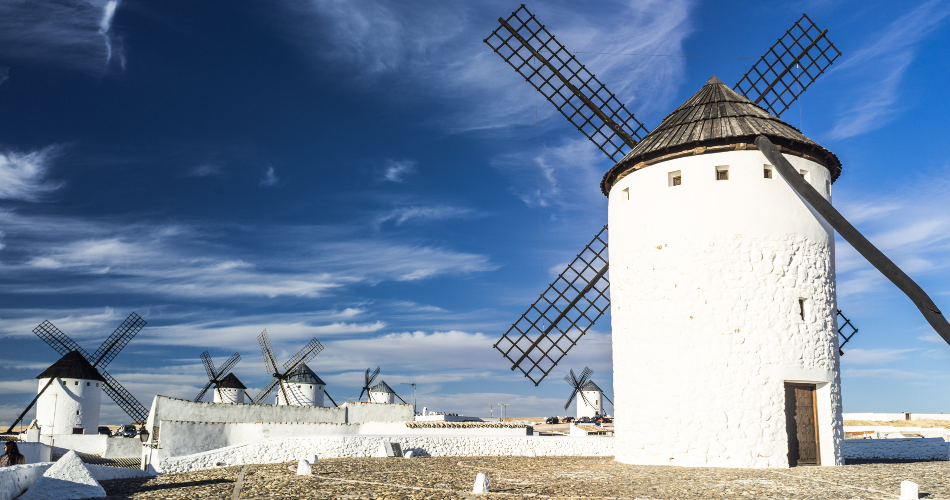 Zdjęcie. Młyny wiatrowe umieszczone blisko siebie. Pojedynczy młyn ma kształt walca, na górze umieszczoną stożkową kopułę, do której przymocowane są po cztery śmigi, zbudowane z kratki drewnianej. Każdy młyn posiada również belkę przymocowaną do kopuły, umożliwiającą obrót nią.