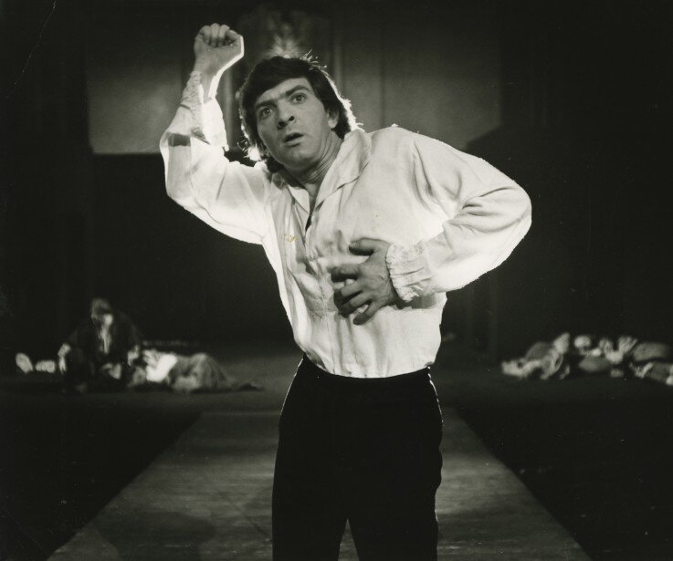 Zdjęcie przedstawia mężczyznę stojącego na scenie. Aktor ma owalną twarz. Czoło postaci jest zasłonięte dość długą grzywką. Mężczyzna ma krzaczaste, zmarszczone brwi. Jego oczy są duże. Postać patrzy przed siebie. Mężczyzna ma gładko ogolone policzki i otwarte usta. Jego włosy są ciemne, proste i sięgające za uszy. Aktor unosi prawą rękę. Lewą dłoń trzyma na sercu. Jest ubrany w jasną koszulę i ciemne, wąskie spodnie. W tle ukazane są inne postaci.