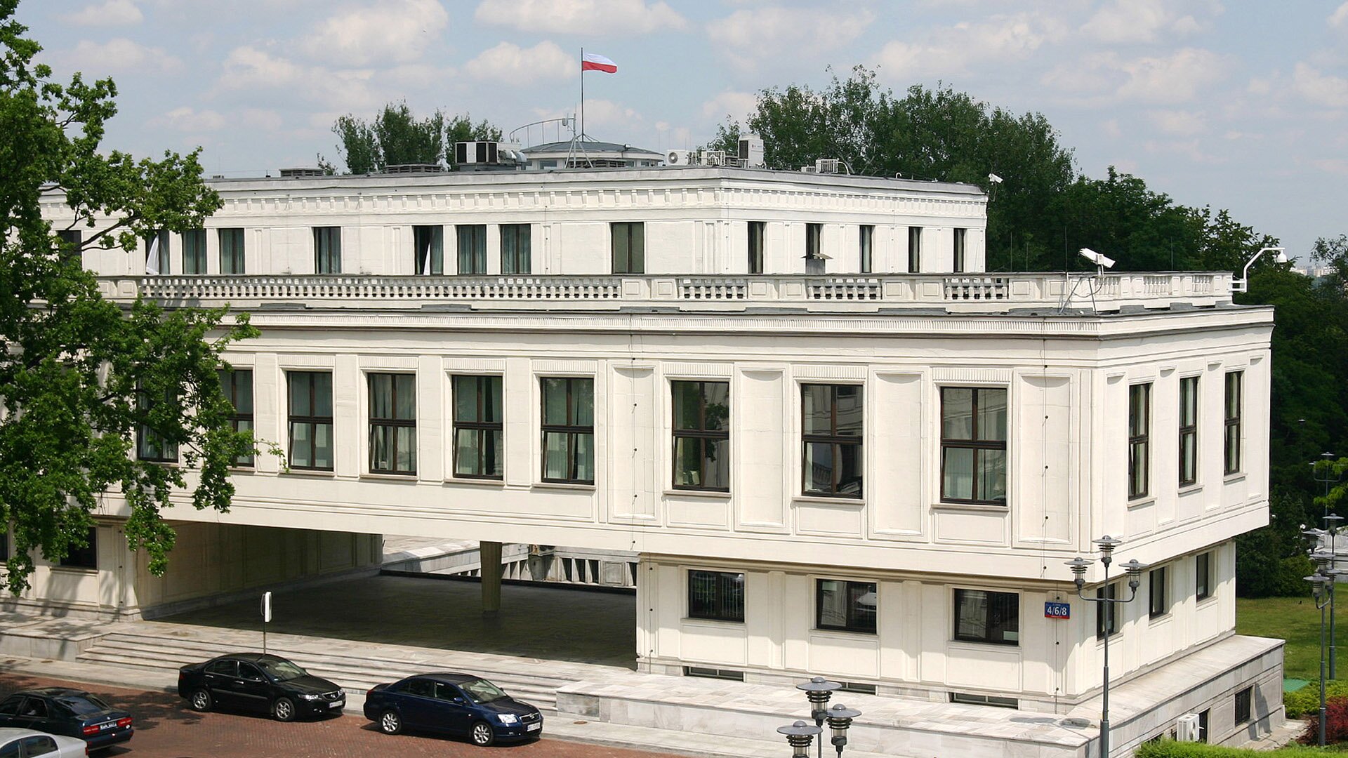 Fotografia przedstawiająca budynek Senatu Rzeczypospolitej Polskiej. Ma trzy poziomy. Drugie piętro zwieńczono biało-czerwoną flagą. Budynek jest jasny, stoi na podwyższeniu.