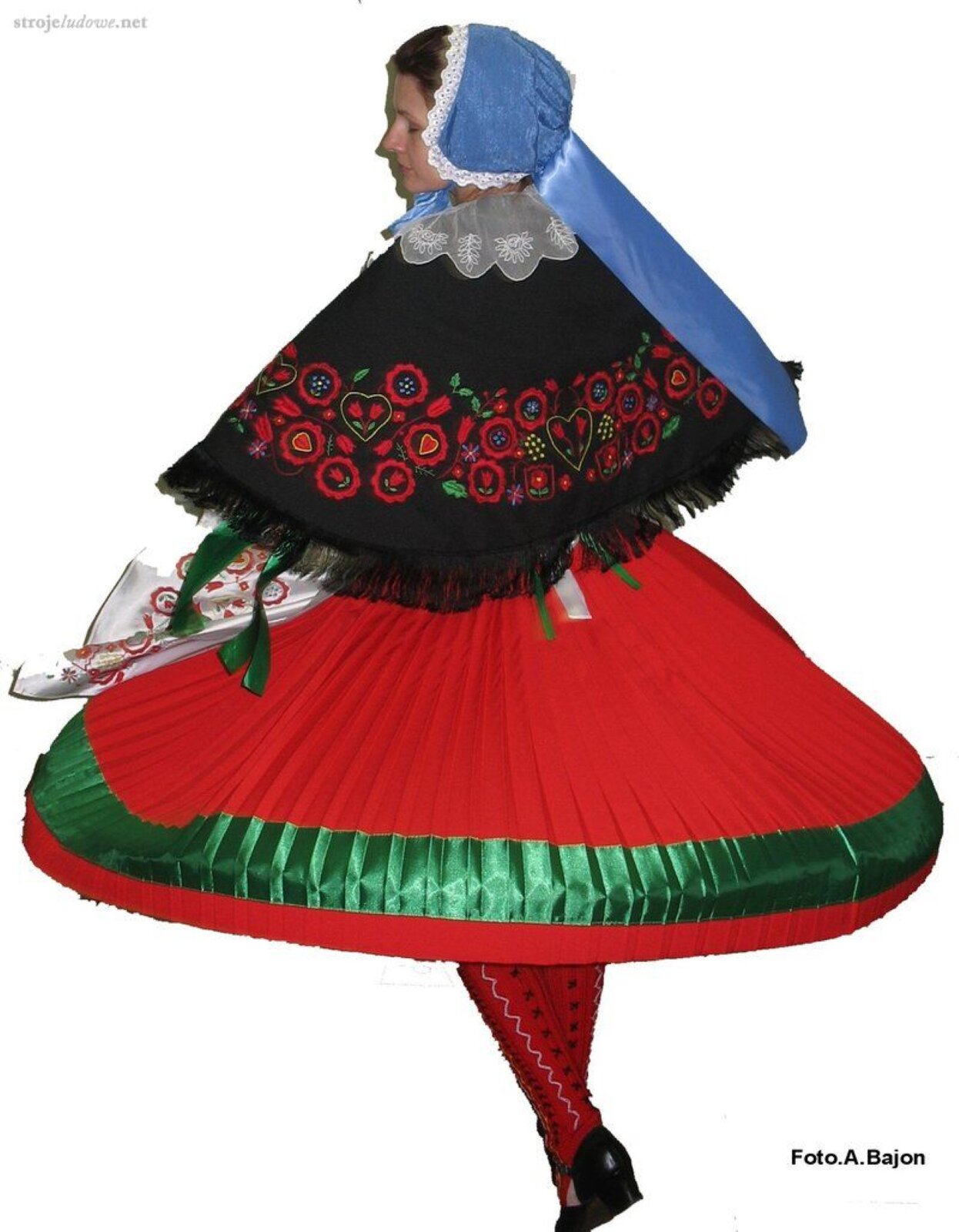 Na zdjęciu znajduje się tancerka w stroju ludowym. Kobieta od dołu ma na sobie następujące części garderoby: czarne, niskie półbuty; haftowane czerwone pończochy; czerwona, plisowana spódnica z zieloną wstążką u dołu; długie, zielone wstążki na rękawach; biały fartuszek; czarna chusta z czerwonymi, haftowanymi kwiatami; biała kryza na szyi.