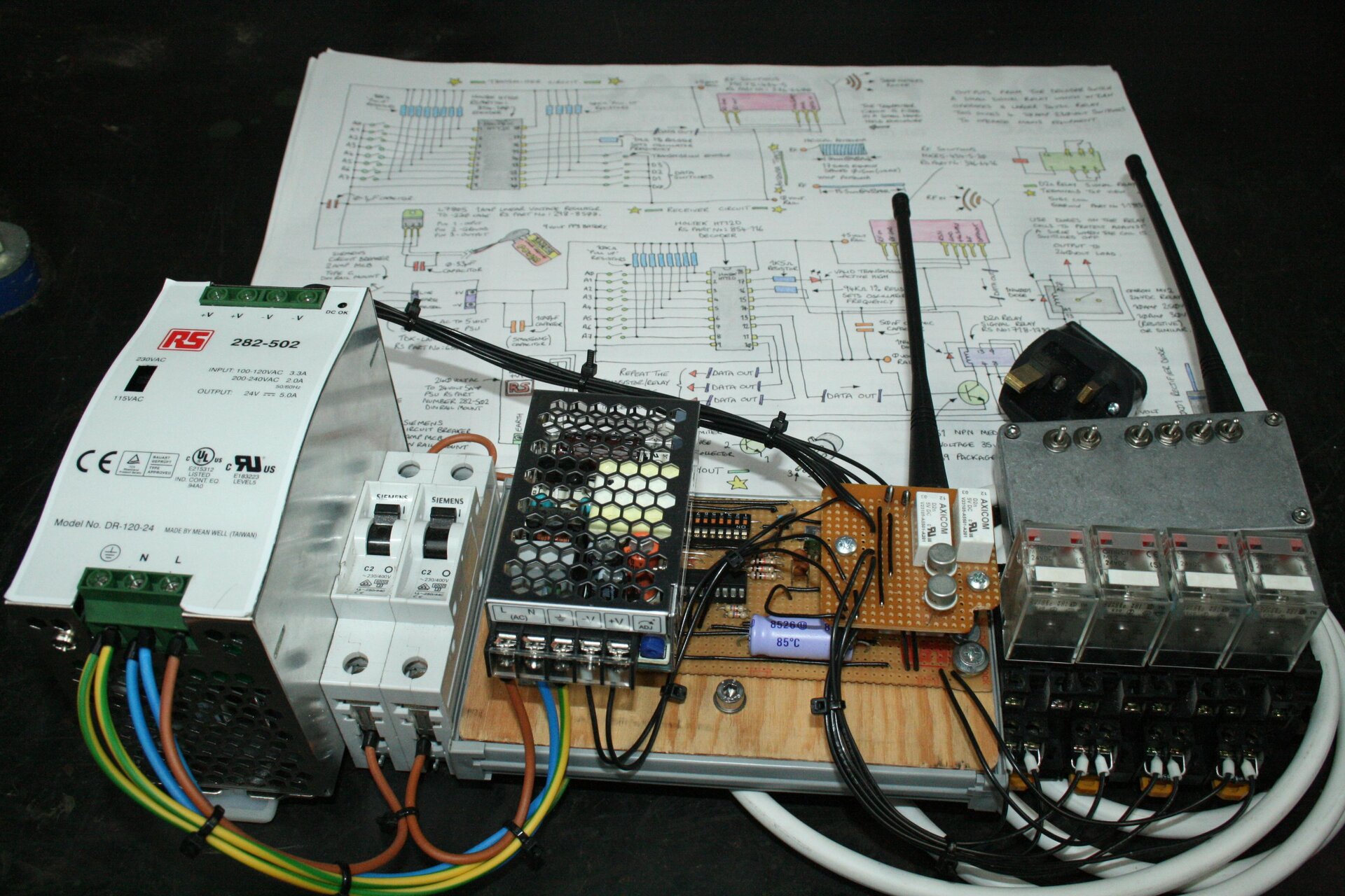 Zdjęcie przedstawia urządzenie elektroniczne domowej roboty składające się z kilku modułów połączonych ze sobą różnokolorowymi przewodami zamontowanych na metalowo drewnianej ramie. Uwagę zwraca obecność dwóch anten świadczących o tym, że jest to jakiś rodzaj nadajnika. Urządzenie leży na pliku kartek, z których pierwsza, najlepiej widoczna jest narysowanym odręcznie schematem. Tło zdjęcia jest czarne.