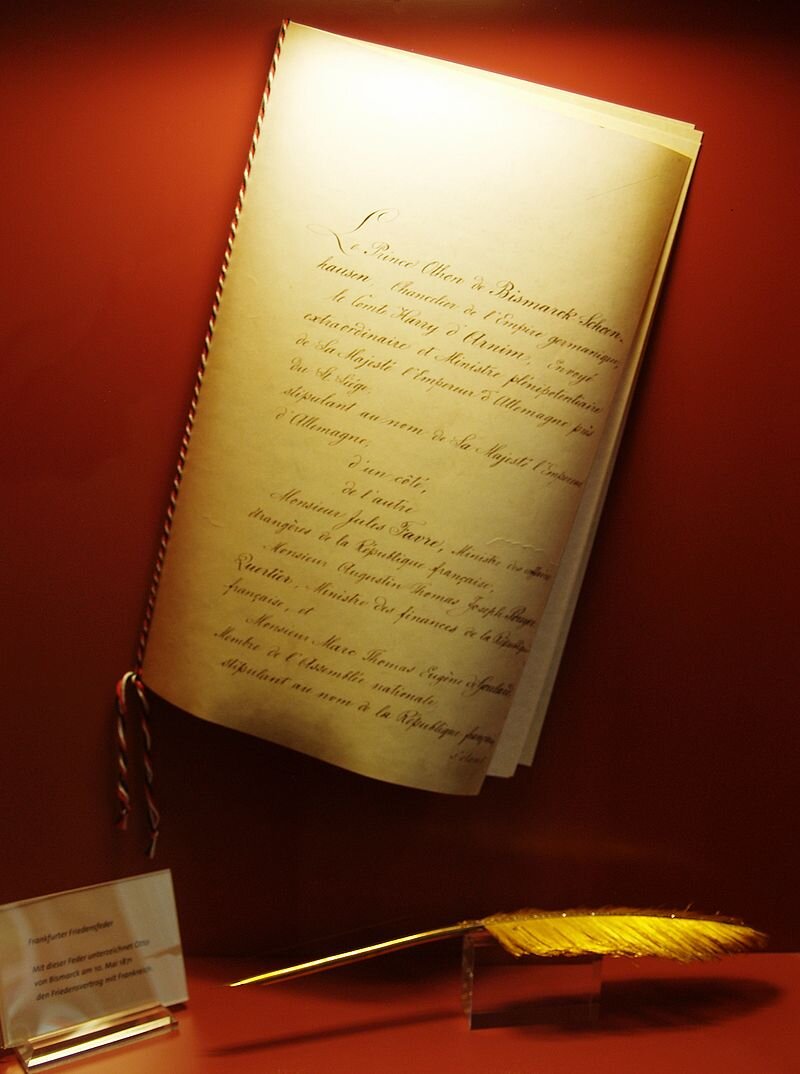 Zdjęcie przedstawia zapisaną ręcznie stronę, która znajduje się w szklanej gablocie. Pod nią są żółte pióro oraz tabliczka z napisem.