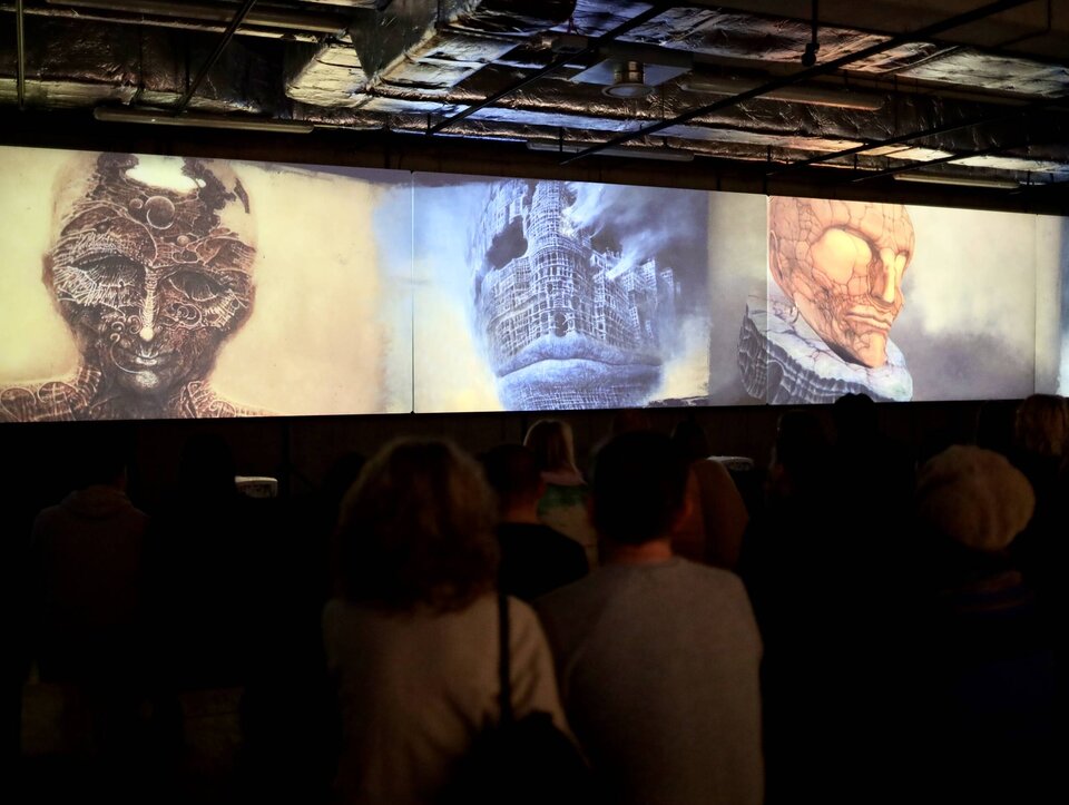 Ilustracja przedstawia kilka osób oglądających prace Zdzisława Beksińskiego na nocnej wystawie. Osoby widzą trzy dzieła artysty - wszystkie przedstawiają wizerunki osób jako surrealistyczne wizje przyszłości, a ich twarze przypominają ruiny lub konstrukcje przypominające maszyny, postacie wydają się być uwięzione w bezlitosnym świecie. Obrazy mogą wywoływać uczucia niepokoju i niepewności, wykazują się także niezwykłą wyobraźnią, techniczną precyzją i bogactwem detali. Postać na obrazie z lewej strony jest barwy brązowej, po środku – niebieskiej, a po prawej stronie pomarańczowej. Na suficie pomieszczenia, w którym miejsce ma wystawa znajdują się duże rury systemu wentylacyjnego.