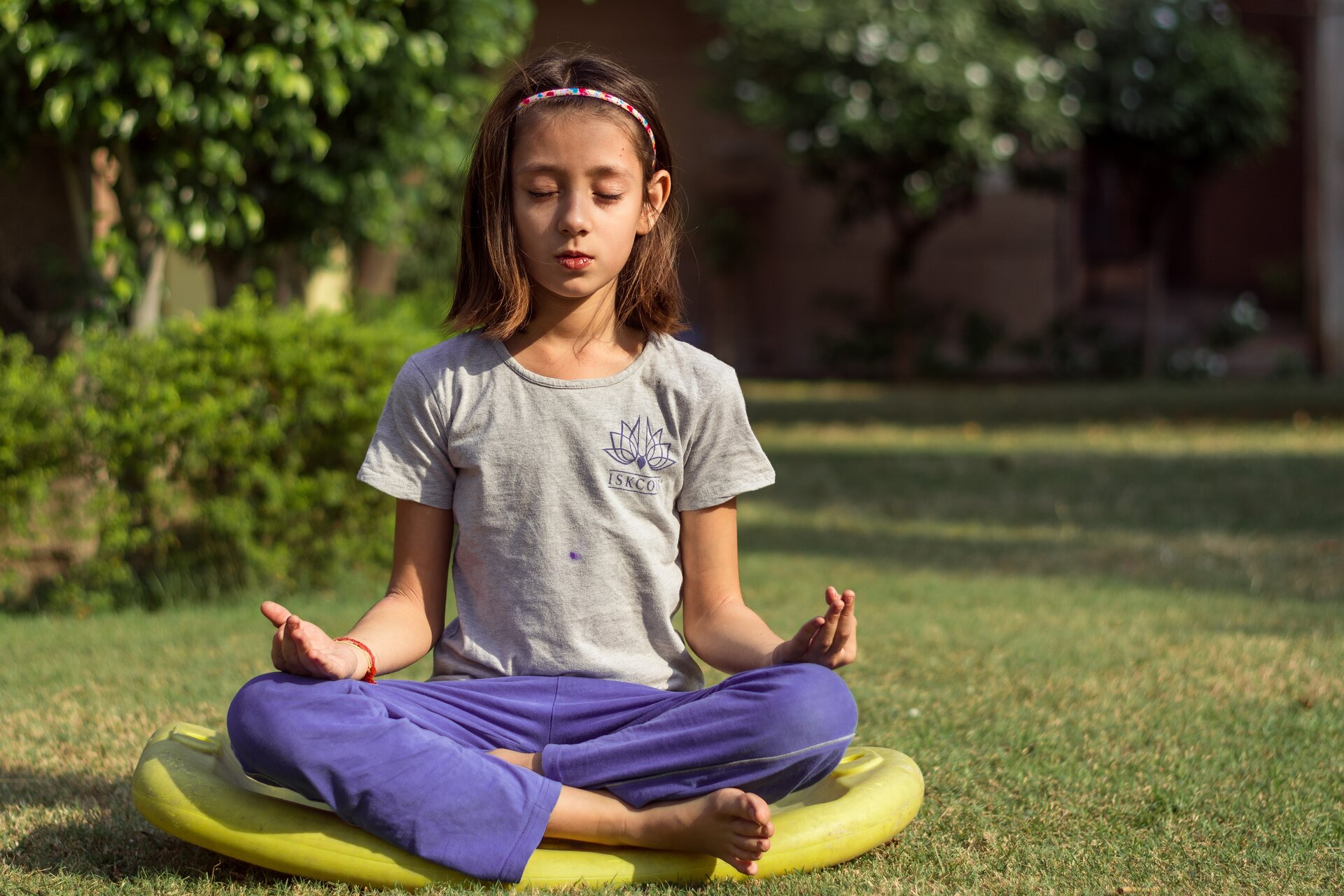 Fotografia barwna przedstawiająca około 11-letną dziewczynkę siedzącą po turecku na macie w ogrodzie. Mata leży na krótko przyciętym trawniku. Jest ciepło, dziewczynka ma na sobie t-shirt z krótkim rękawem i cienkie długie spodnie. Ma bose stopy. Oczy ma zamknięte, a ręce rozłożone, dłonie oparte na kolanach. Dziewczynka praktykuje jogę. W tle widać drzewa i krzewy oraz niewyraźne zabudowania.