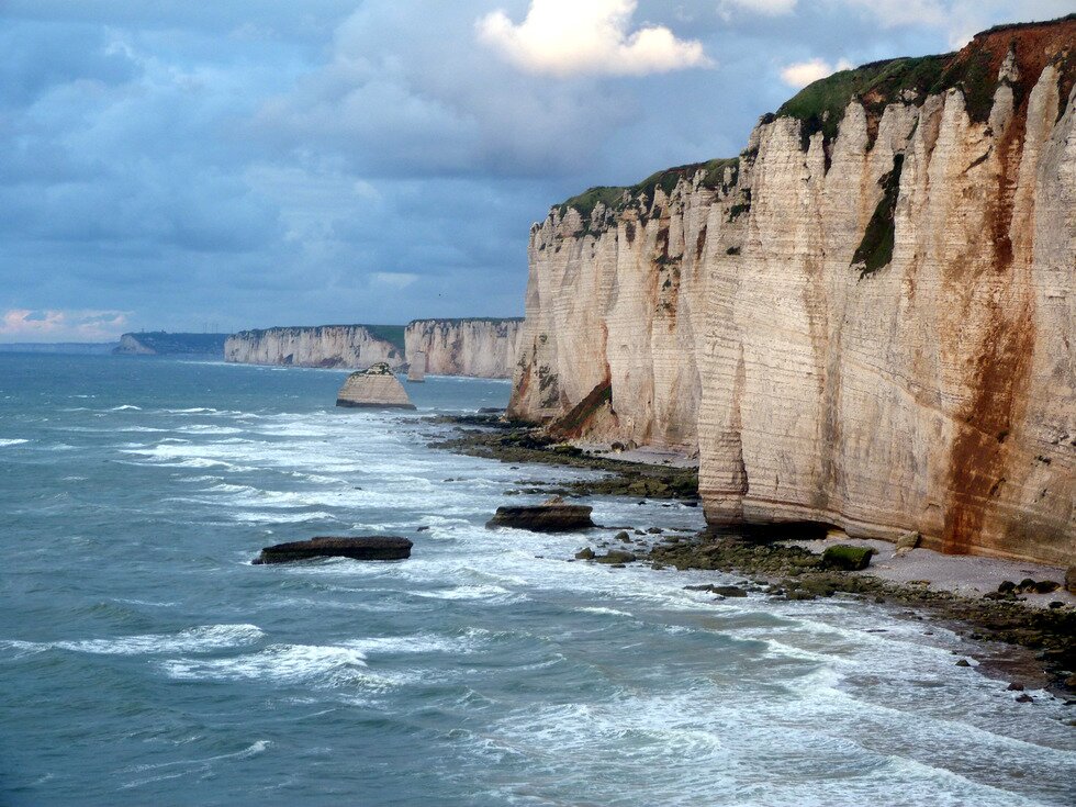 Na zdjęciu nadmorski klif. Wysoka, bardzo stroma ściana skalna. U jej podstawy wąska plaża. Niebieskie morze. Białe fale.
