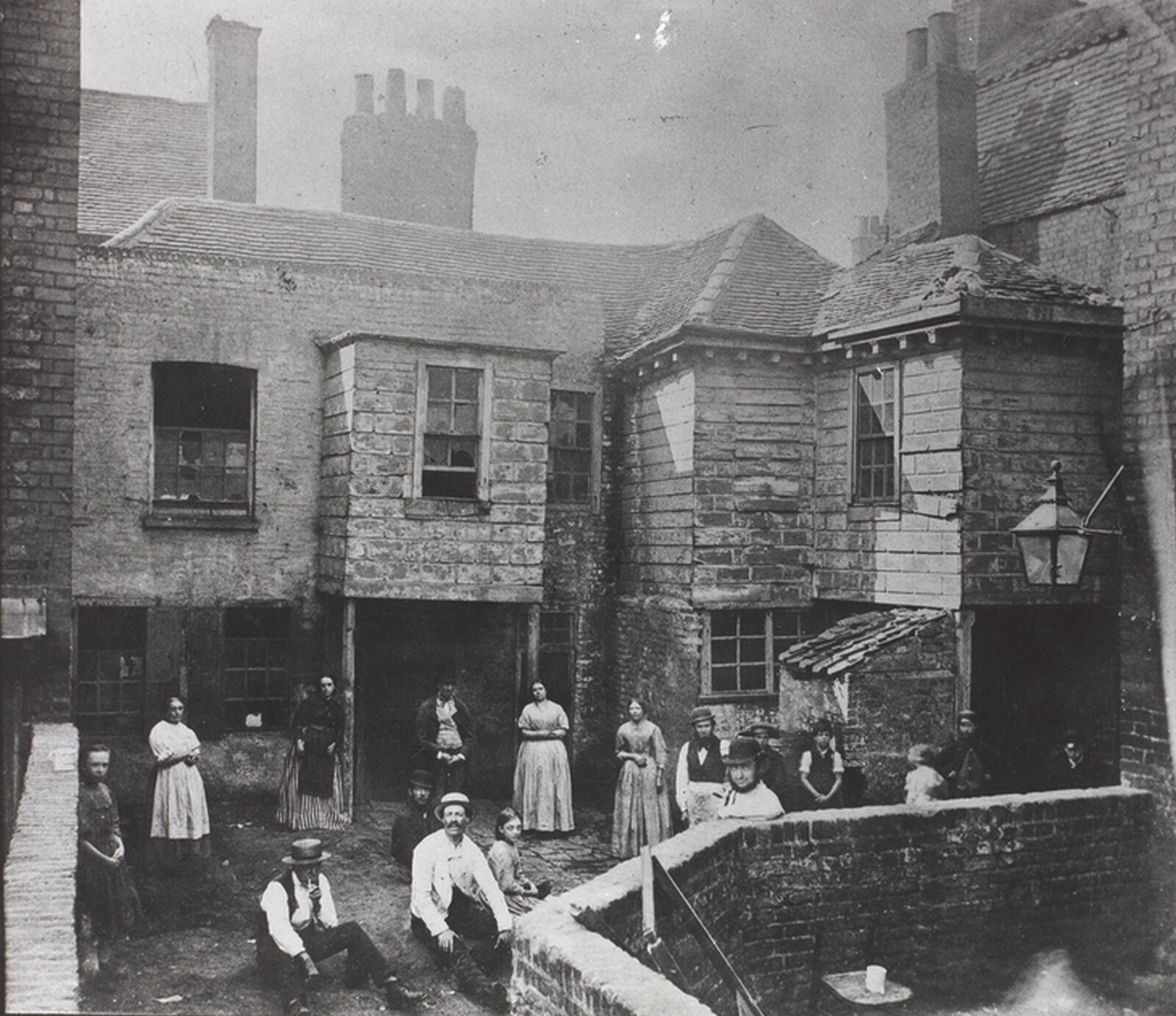 Stara czarnobiała fotografia niewielkiego, ceglanego budynku o nieregularnym kształcie. Przed nim bardzo małe podwórko, otoczony innymi budynkami. Na podwórku grupa osób - mężczyzn, kobiet i dzieci. Część osób stoi, a część siedzi na ziemi. Kobiety ubrane w długie proste suknie, mężczyźni w jasne koszule i ciemne spodnie.