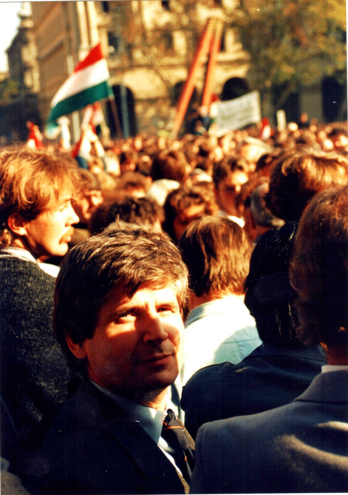 Zdjęcie przedstawia tłum manifestujących ludzi na ulicy. Na pierwszym planie w stronę widza patrzy młody mężczyzna. Pozostali uczestnicy protestu są skierowani w drugą stronę. Trzymają flagi Węgier oraz transparenty. W tle widoczny jest budynek oraz drzewo.