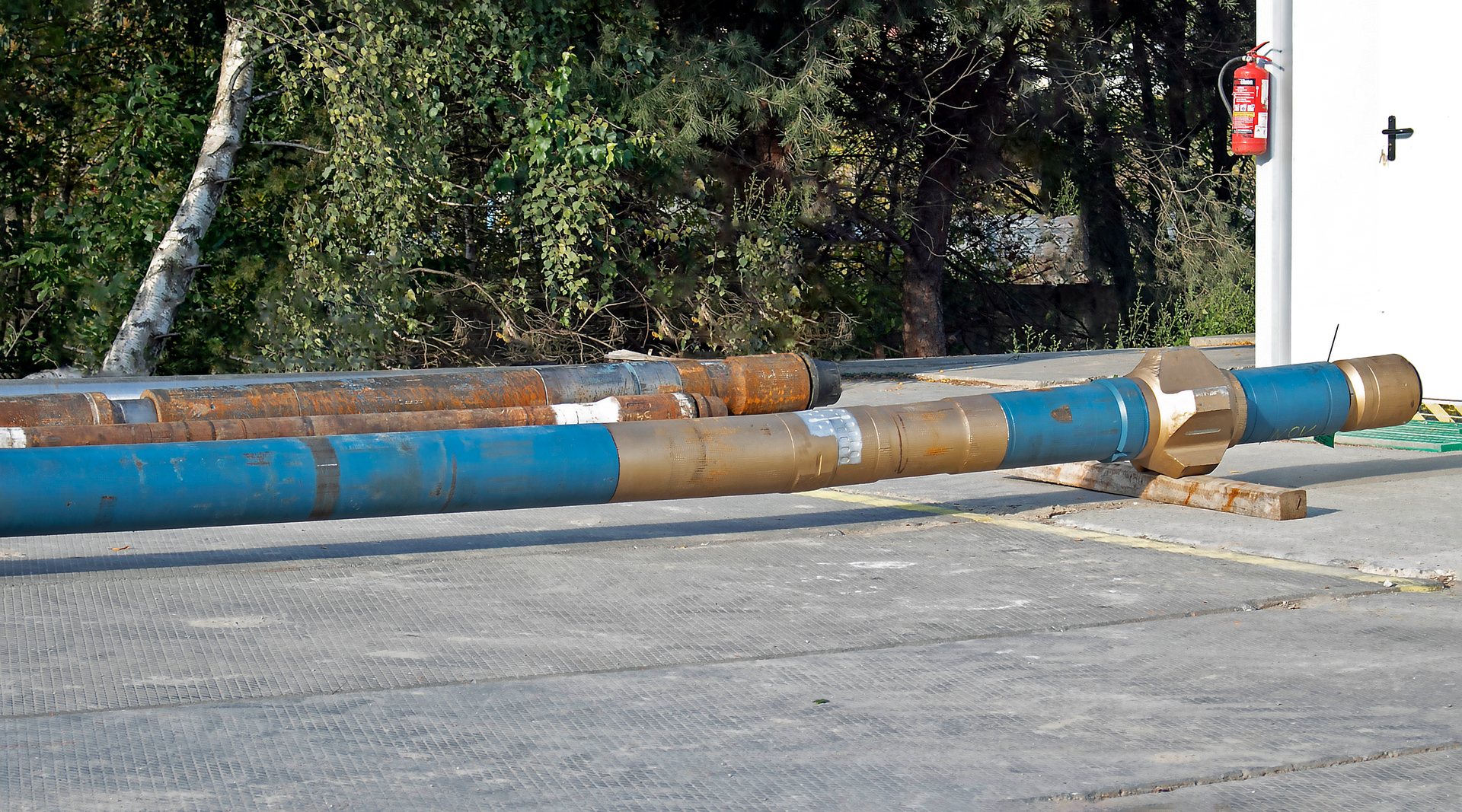 Zdjęcie przedstawia krzywy łącznik. Jest on zorientowany poziomo na placu wiertni. W tle znajdują się drzewa. Krzywy łącznik zamontowany jest między silnikiem wgłębnym a świdrem wiertniczym. Stanowi go rurowaty element wykonany z brązowawego, lekko połyskującego metalu. Z lewej strony odchodzi od niego niebieska rura. Podobnie z prawej. Przy czym jest ona tutaj zdecydowanie krótsza i nagwintowana jest na cześć składającą się z czterech symetrycznych względem osi poprzecznej i rozmieszczonych wokół obwodu rury wypustek.