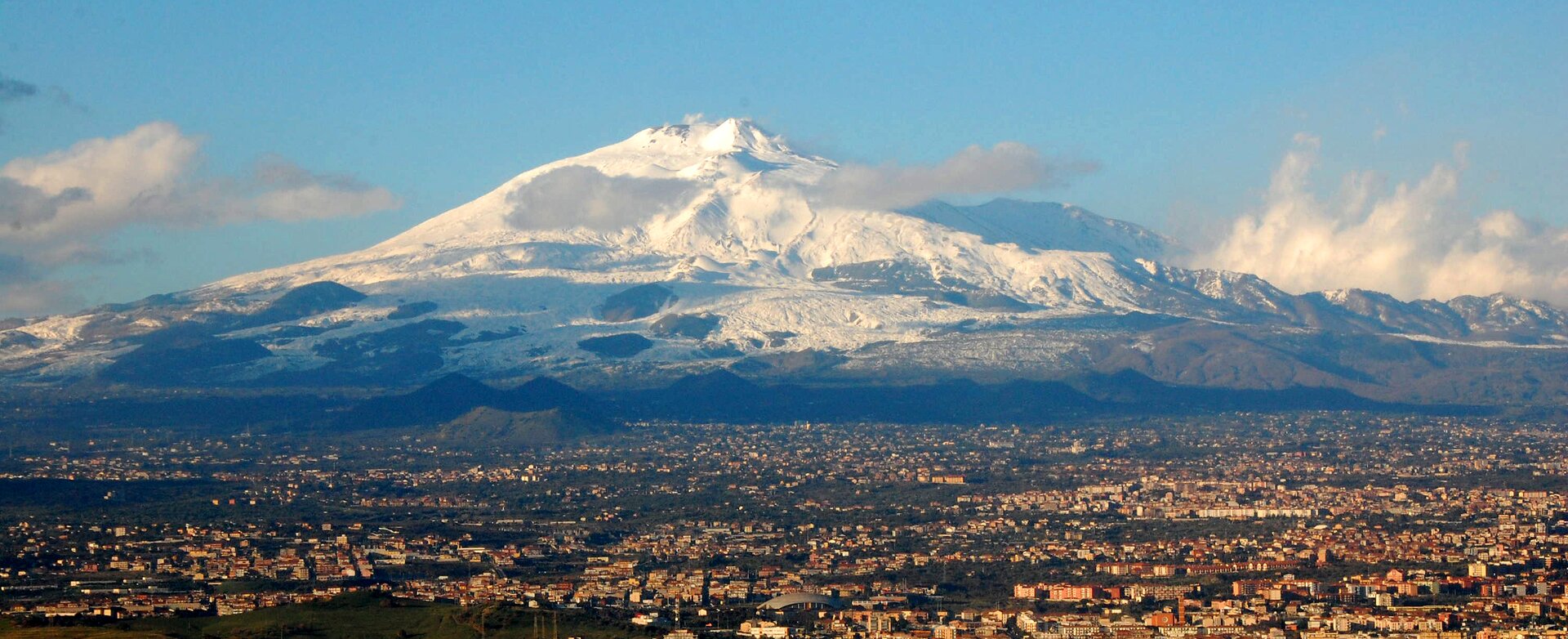 Na zdjęciu duży teren zabudowany w tle znajduje się duży wulkan Etna, nad nim niebieskie niebo z kilkoma chmurami. 