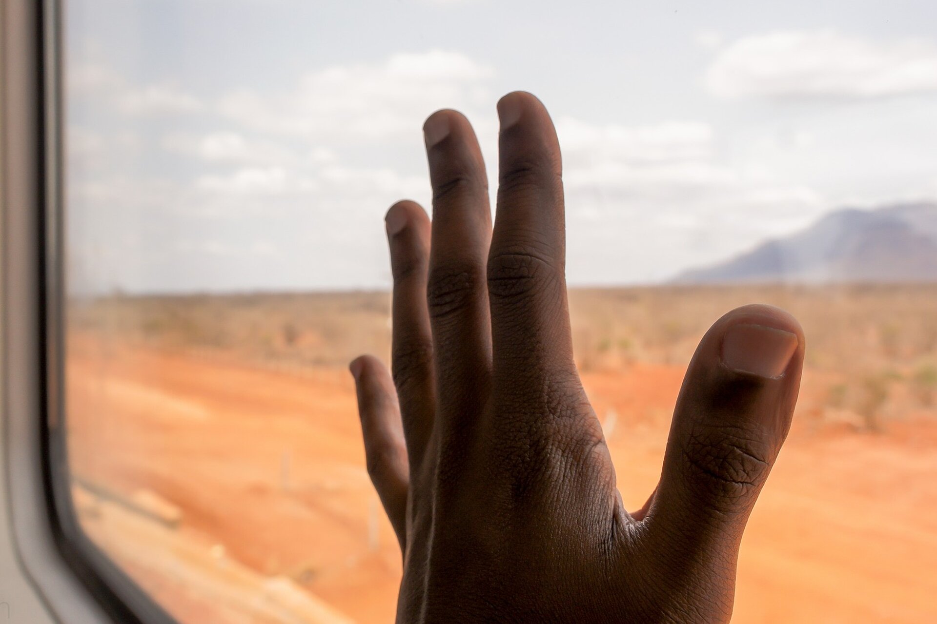 Zdjęcie przedstawia dłoń położoną na szybie okna pociągu. Za oknem widoczna jest rozległa równina, a w oddali masyw górski.