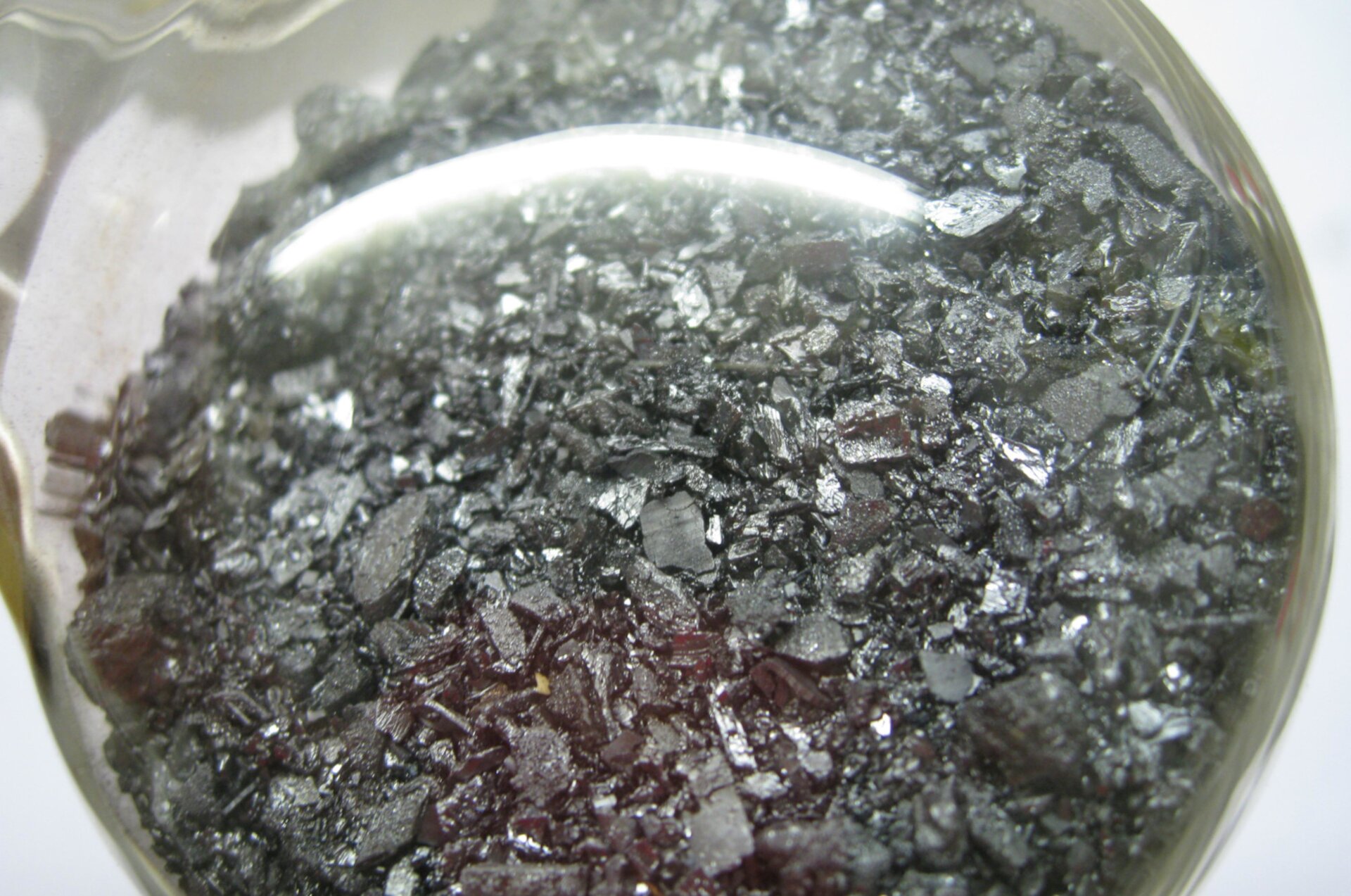 Na ilustracji ukazano zbliżenie na dolną część kolby okrągłodennej, w której znajduje się wiele drobnych kawałków ciemnej, połyskującej skały.