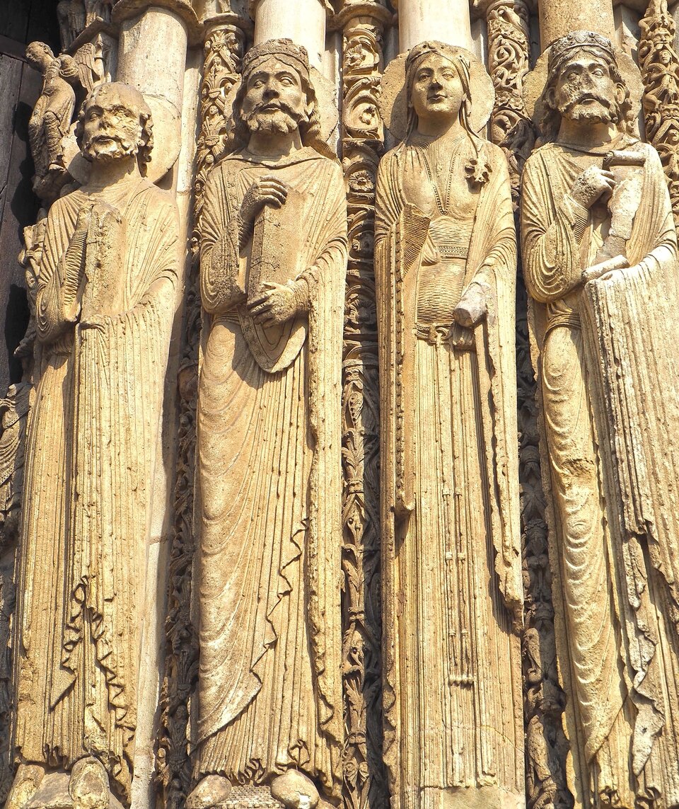 Ilustracja przedstawia cztery rzeźby figuralne z zachodniego portalu katedry w Chartres. Są podniosłe, sztywne, nieruchome, koloru piaskowego. Mają wzrok skierowany przed siebie. W rękach trzymają zwoje lub księgi. Ich szaty opadają swobodnie. Na twarzach pojawia się delikatny uśmiech.