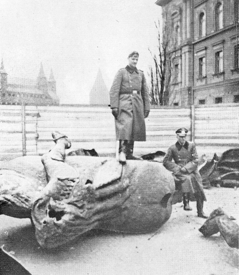 Zdjęcie przedstawia dwóch niemieckich żołnierzy. Jeden stoi na elemencie pomnika, drugi siedzi. W tle wieże Krakowa.