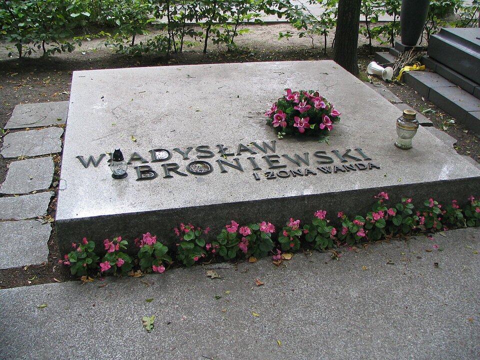 Zdjęcie przedstawia kamienną, kwadratową płytę nagrobną z napisem: Władysław Broniewski i żona Wanda. Na płycie stoją dwa znicze oraz leży mały wieniec z fioletowych kwiatów. Przed płytą rośnie rząd drobnych, różowych kwiatków. Nagrobek otoczony jest chodnikiem ułożonym z kamiennych płyt. W tle widać pień drzewa i krzewy. Po prawej stronie - kolejny nagrobek, a obok niego przewrócony wazon i suche kwiaty.
