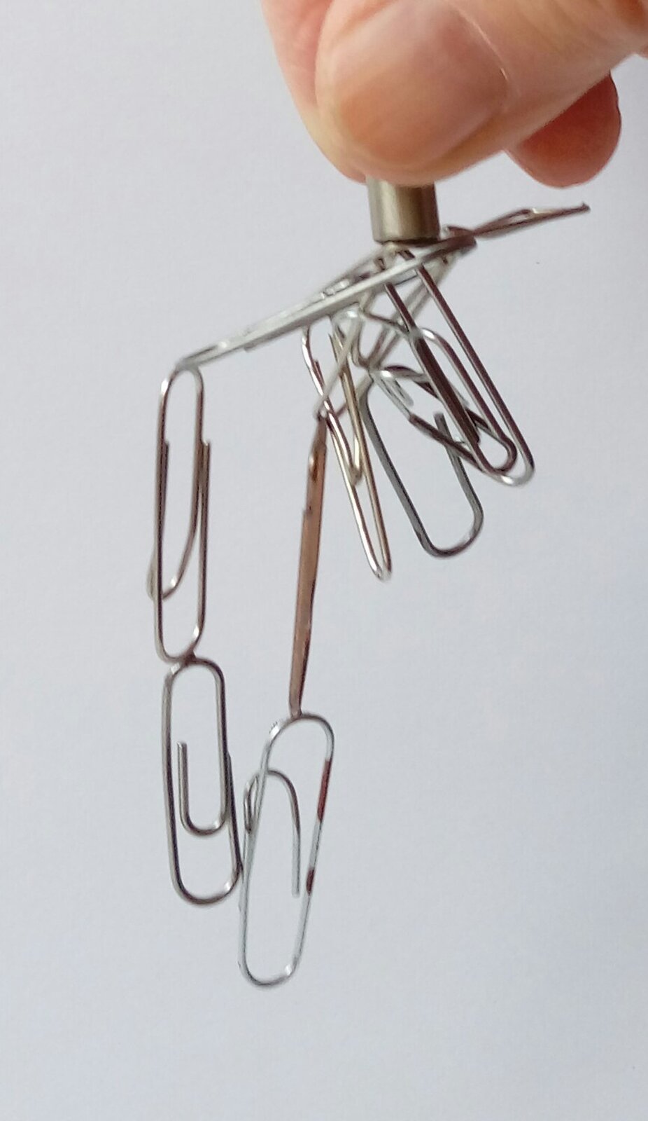 Zdjęcie przedstawia palce dłoni (kciuk i palec wskazujący) trzymające magnes, do którego przyczepionych jest wiele spinaczy biurowych (spinacze pod wpływem magnesu przyciągają się wzajemnie).