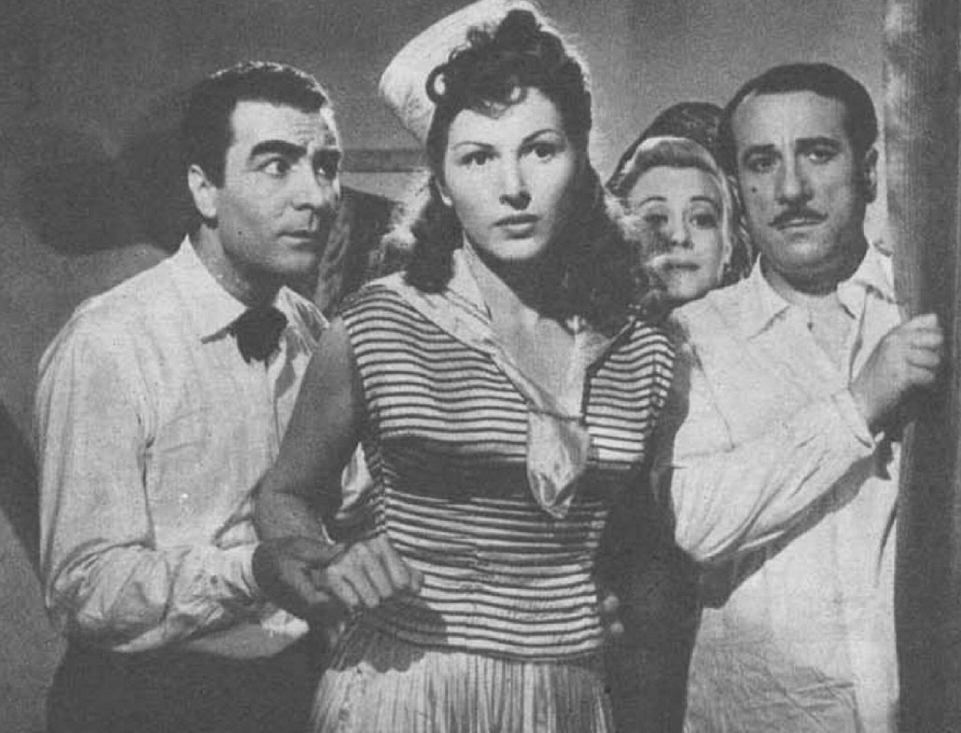 Zdjęcie przedstawia kadr z filmu "Światła variété" reżyser Federico Fellini. Cztery postacie, dwie kobiety i dwaj mężczyźni pokazani są od pasa w górę. Między dwoma mężczyznami stoi kobieta ubrana w marynarską bluzę, na głowie ma czapkę majtka. Po prawej stronie między kobietą a mężczyzną widać twarz innej kobiety.