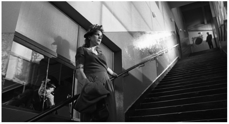Zdjęcie przedstawia wysokie schody w fabryce. Przy dyżurce pilnowanej przez portiera stoi młoda kobieta w letniej sukience i kapeluszy. Czeka. Na szczycie schodów znajduje się postać mężczyzny w garniturze. Nie widać jego twarzy.