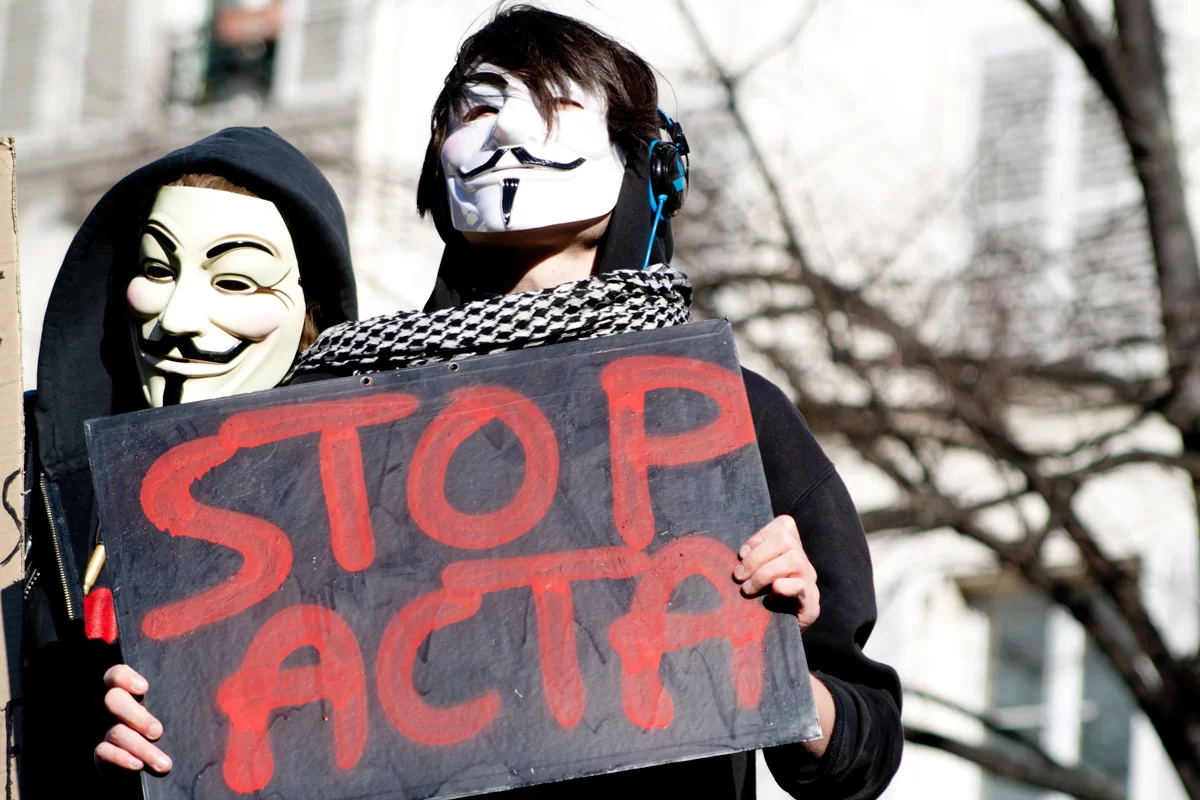Zdjęcie przedstawia dwie osoby w maskach z uśmiechem. Jedna z nich trzyma karton z napisem: Stop Acta.
