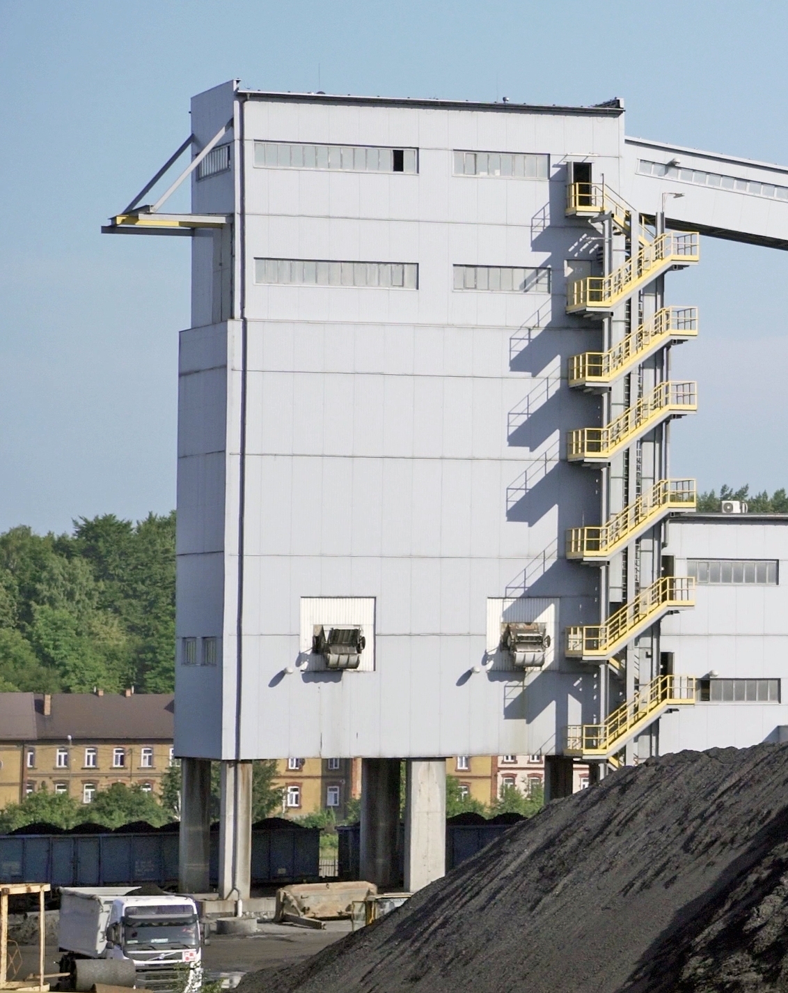 Na zdjęciu widoczny jest budynek magazynowy. Budynek jest obiektem wielkopowierzchniowym o kształcie prostopadłościanu. Po prawej stronie budynku przylega do niego klatka schodowa. W górnej części budynku znajduje się łącznik. Łącznik jest nachylony względem gruntu.Przed obiektem widać hałdę węgla. Obiekt posadowiony jest na prostopadłościennych filarach. Pomiędzy filarami widać wagony załadowane węglemPoniżej zdjęcia widoczny jest przycisk multimedialny z nagraniem audio. Treść nagrania jest zgodna z tytułem zdjęcia.