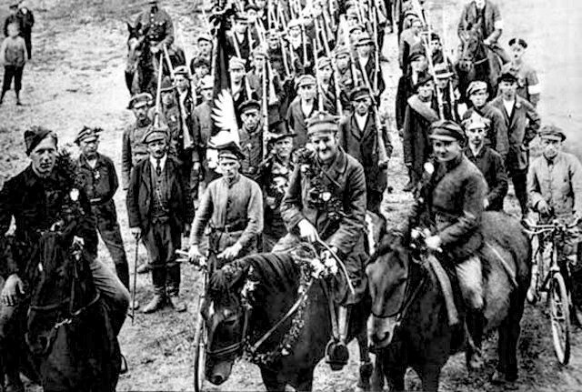 Fotografia przedstawia oddział powstańczy. Prości chłopi zagarnięci do walki. Niektórzy z rowerami, w zwykłym codziennym ubiorze z szarfą na ramieniu. Każdy przerzuca przez ramię strzelbę. Przed nimi oficerowie konno. 