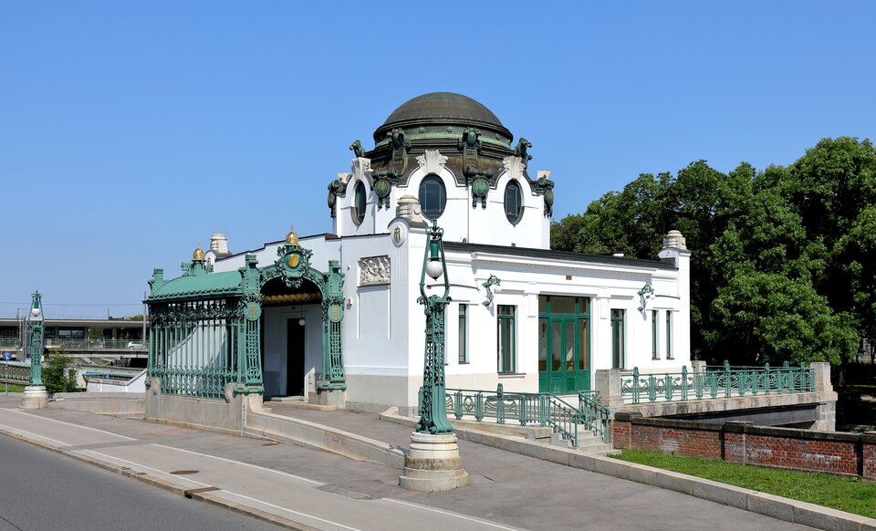Secesyjny pawilon Hofpavillon Hietzing w Wiedniu, usytuowany przy ulicy i otoczony zielenią. Biały budynek ma kopułę z ciemnej patynowanej miedzi, ozdobioną dekoracjami i okrągłymi oknami. Elewację zdobią zielone metalowe ornamenty, stylizowane motywy roślinne oraz dekoracyjne latarnie. Po lewej stronie znajduje się przeszklony, bogato ornamentowany przedsionek wejściowy. Przed budynkiem biegnie chodnik i jezdnia. W tle po prawej widoczne są drzewa, a po lewej fragment infrastruktury kolejowej. Nad budynkiem rozciąga się bezchmurne, błękitne niebo.