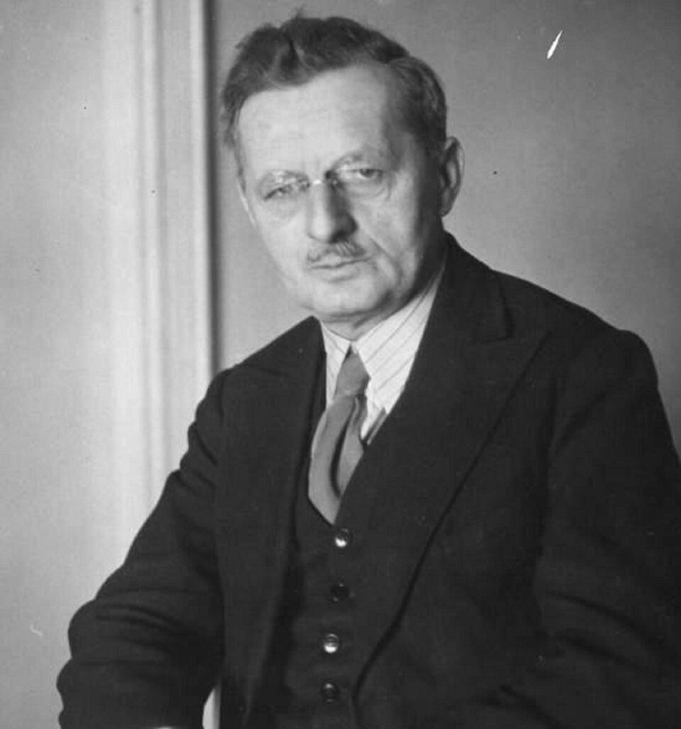 Ilustracja przedstawia portret Ludomira Michała Rogowskiego - polskiego kompozytora i dyrygenta. Mężczyzna z poważną miną w okularach bez oprawek i zauszników. Ma delikatne wąsy i bardzo poważną minę. Ubrany jest w białą koszulę, szary krawat i garnitur.