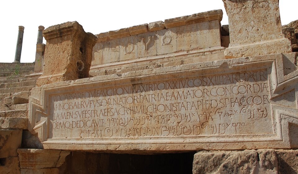 Zdjęcie przedstawia fragment zabytku architektonicznego - tablicę z 5 wersami napisu. Nad nimi umieszczono płytę z piaskowca z widocznymi dwoma wieńcami laurowymi. W górnej lewej części ilustracji widoczny fragment antycznej kolumnady, a także stopnie, ułożone poniżej schodkowo. Zdjęcie wykonane w jasny dzień. 