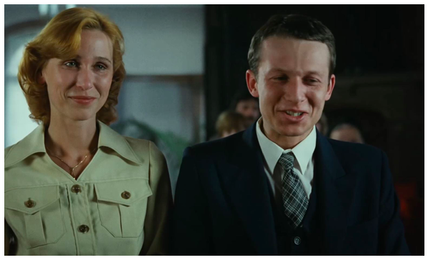 Fotografia przedstawia kobietę i mężczyznę. Są to ludzie w sile wieku. Kobieta stoi po lewej stronie. Jej twarz jest owalna, ukazana w całości. Postać ma gładkie czoło. Jej brwi są cienkie i ciemne. Ma głęboko osadzone oczy i prosty nos. Usta kobiety są wydatne, wokół nich zarysowują się zmarszczki mimiczne. Włosy lekko falują, są jasne i sięgają szyi. Jest ubrana w żakiet z kołnierzem i kieszeniami na piersiach. Nosi na szyi cienki, złoty łańcuszek z zawieszką ukrytą pod ubraniem. Mężczyzna, stoi po jej lewej stronie. Ma owalną twarz, którą prezentuje w całości. Jego czoło jest wysokie i szerokie. Pośrodku czoła odznacza się pionowa żyła. Ma zmarszczone brwi, przymrużone oczy i okazały nos. Spogląda w dół. Wargi są uchylone, odsłaniając zęby. Jego włosy są krótko i starannie ostrzyżone. Ubrany jest w garnitur. Za ich plecami stoi grupa osób.