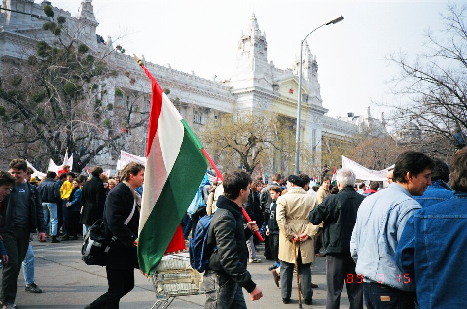 Zdjęcie przedstawia tłum protestujących ludzi na miejskim placu. Trzymają transparenty. Na środku zdjęcia mężczyzna trzyma flagę Węgier. Za manifestującymi znajdują się drzewa oraz duży budynek zwieńczony czerwoną gwiazdą.