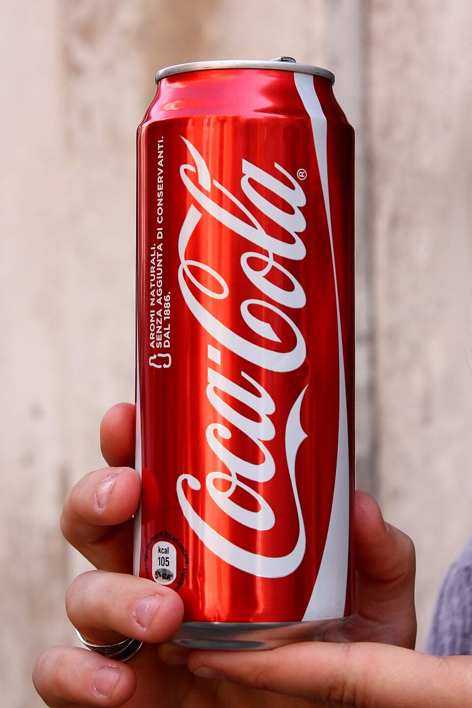 Czerwona puszka napoju Coca‑Cola trzymana w dłoni; na puszce widoczne jest białe logo Coca‑Cola, a w tle znajduje się jasna, drewniana powierzchnia.