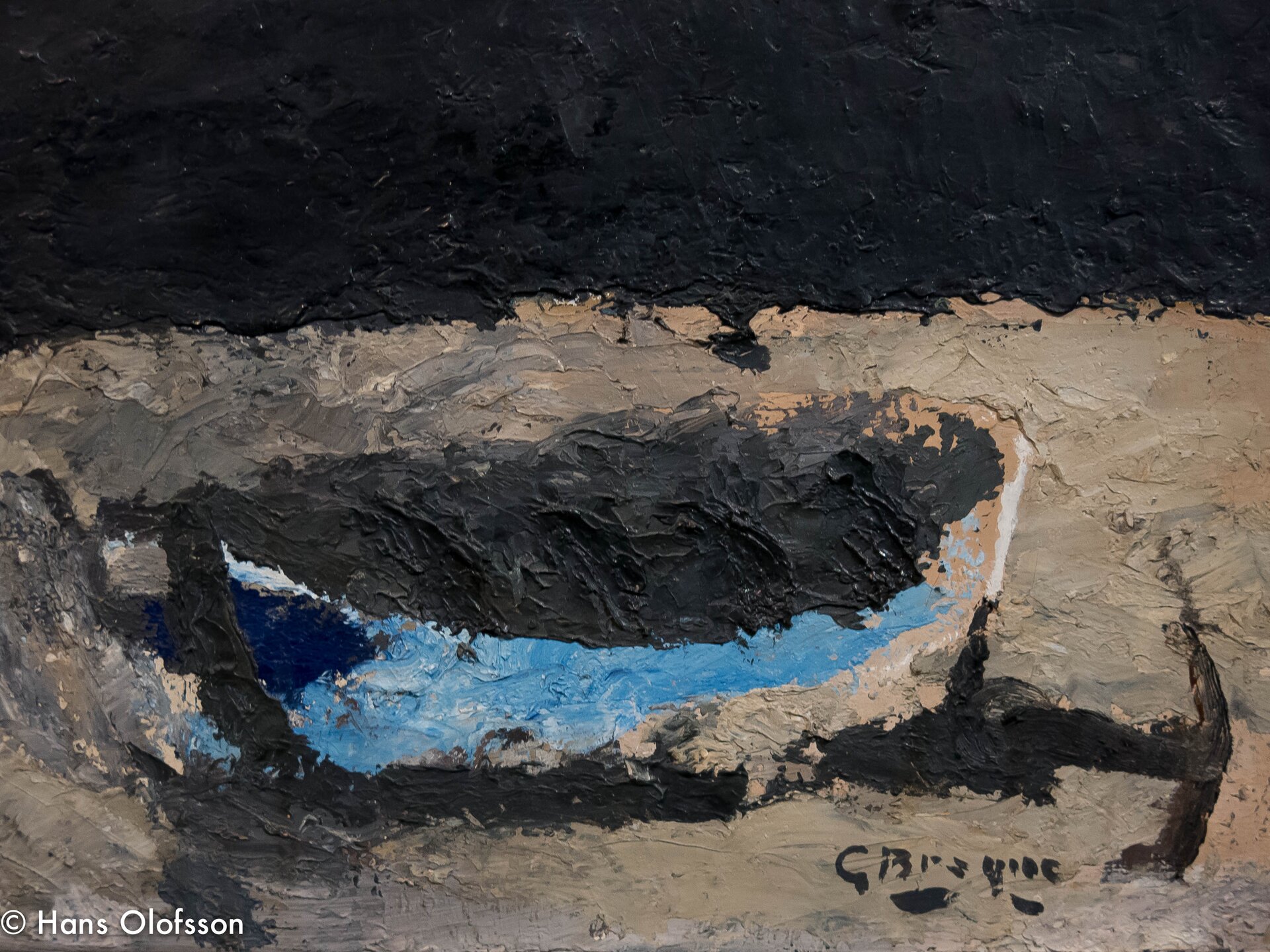 Obraz przestawia niebieską łódkę na plaży. Wnętrze łódki wypełnione grubo nałożoną czarną farbą, która tworzy nieregularną, ostrą fakturę. Plaża namalowana jest gładkimi pociągnięciami pędzla. Morze namalowano czarną farbą, grubo nałożoną.