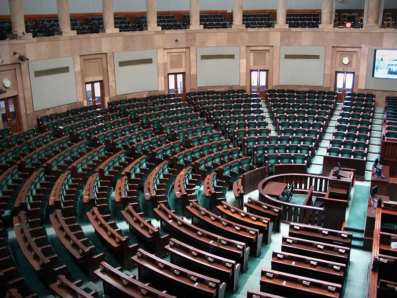 Kliknij, aby powiększyć. Ilustracja przedstawia zdjęcie owalnej sali wypełnionej drewnianymi fotelami obitymi zielonym materiałem, tworzącymi półkole. W centrum znajduje się mównica, a nad nią podwyższenie. Przed mównicą stoją krzesła i stół otoczone półkolistą barierką. W górnej części na balkonach znajdują się miejsca dla widzów.