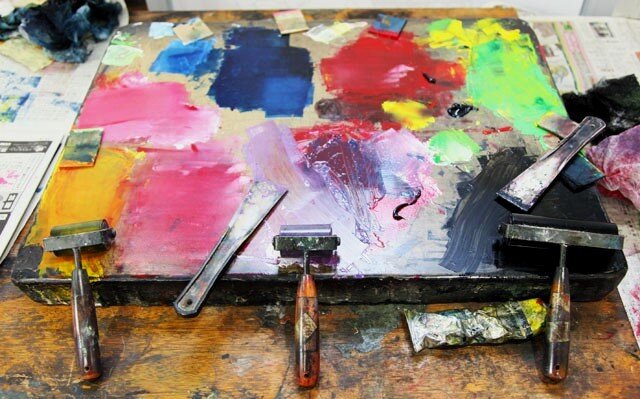  Zdjęcie przedstawia fragment stołu w pracowni malarza, przykrytego w górnej części gazetami, na którym leży metalowa płyta pokryta kolorowymi plamami po farbie. O płytę oparto trzy wałki do rozprowadzania farby, przy jednym leży tubka farby. Z prawej i lewej strony leżą również na płycie dwie szpachle. Płyta pokryta kolorowym plamami farby w kształcie kwadratów i prostokątów - śladów po próbach nakładania wałkiem farby.