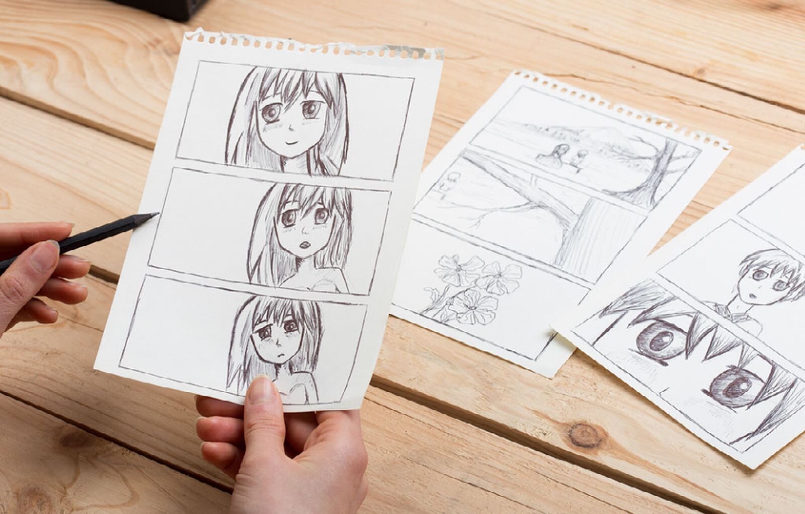 Zdjęcie przedstawia rysownika komiksu. Nad drewnianym stołem pokazane są dłonie rysownika z ołówkiem, trzymające kartkę z rysunkiem dziewczynki. Na rysunku dziewczynka przedstawiona jest w trzech ujęciach, od całej postaci, do portretu. Na stole leżą jeszcze dwie kartki z rysunkami do tego samego komiksu. 