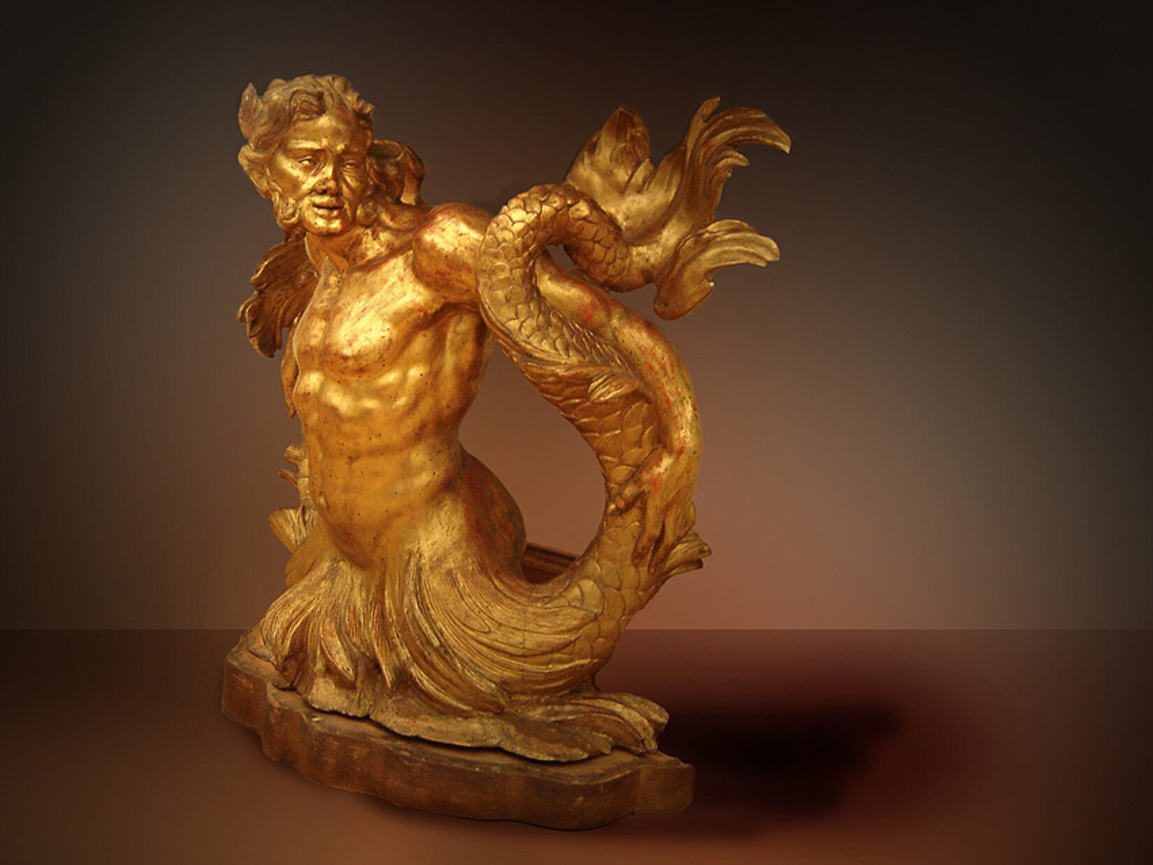 Ukazana na fotografii postać ma tułów człowieka, zaś dół węża. Jest to mitologiczny tryton. Jest mężczyzną w średnim wieku o włosach zaczesanych do tyłu. Ma lekko otwarte oczy oraz usta. Jego ciało jest umięśnione. Lewą ręką podtrzymuje ogon. Rzeźba ma kolor złoty. Umieszczona jest na niskim cokole na tle brązowej oświetlonej ściany.