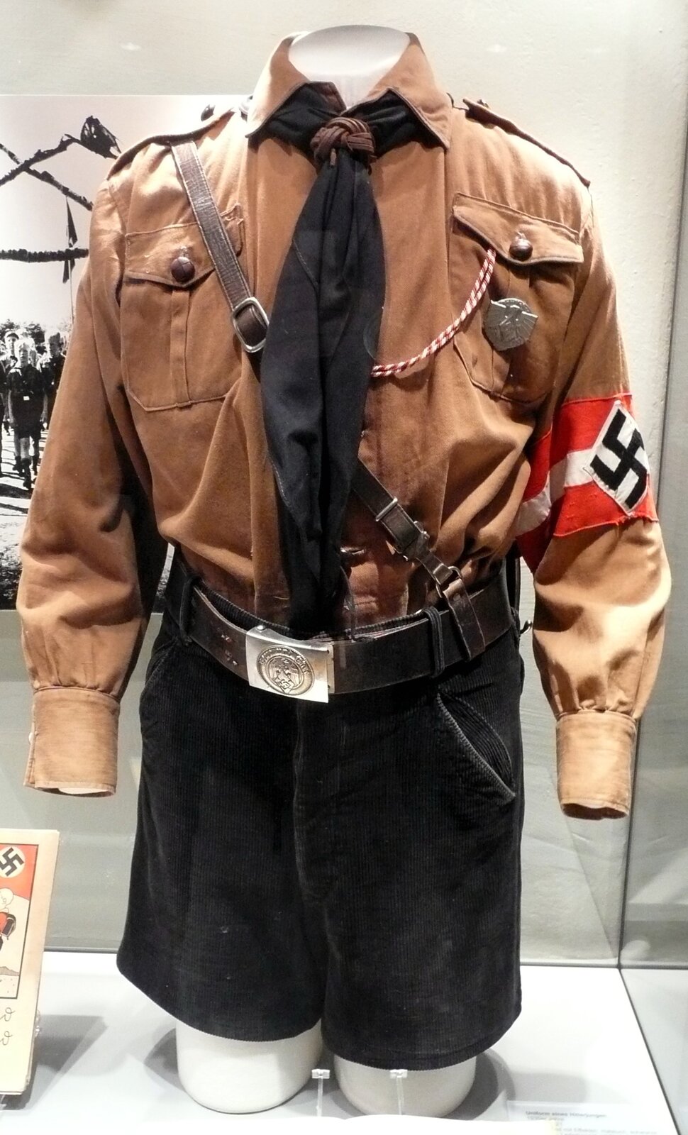Zdjęcie przestawia mundur Hitlerjugend zawieszony na manekinie bez głowy. Mundur składa się z brunatnej koszuli i czarnej chusty. Na lewym ramieniu jest czerwonobiała przepaska ze swastyką. Spodenki są krótkie i czarne, spięte paskiem z klamrą.
