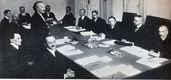 Fotografia przedstawiająca rząd Ignacego Jana Paderewskiego. Jeden z męzczyzna stoi. Ma dłuższe, siwe włosy zaczesane do tyłu, ciemne wąsy. To Ignacy Jan Paderewski. Współpracownicy siedzą przy stole na obradach, mają przed sobą notatki. Wszyscy zgromadzeni ubrani są w ciemne garnitury.