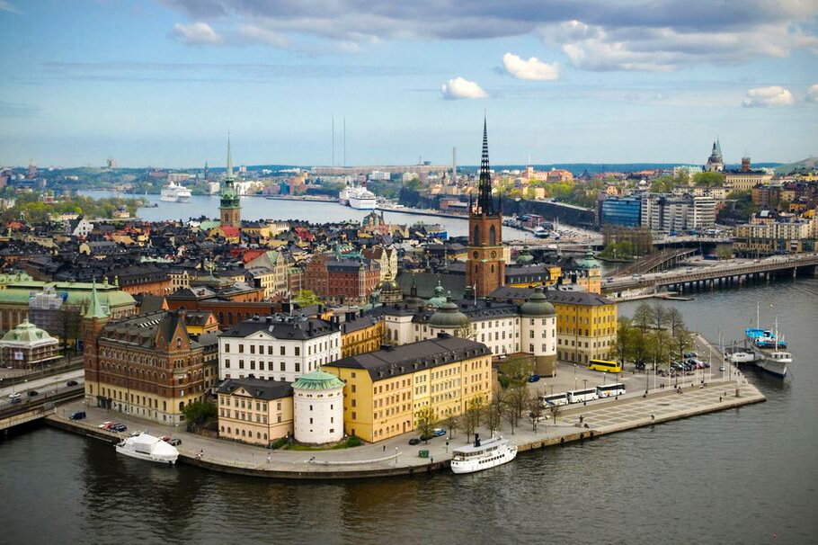 Zdjęcie przedstawia widok z góry na Sztokholm. Budynki mieszkalne, wieże kościołów, mosty, nowoczesne biurowce oraz promy znajdujące się na wodzie.