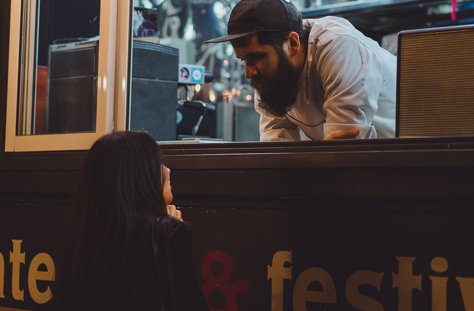 Zdjęcie przedstawia mężczyznę pochylonego i opartego na ladzie food trucka. W tle widoczne jest wyposażenie kuchni samochodu. Poniżej lady widoczna jest kobieta, zwrócona w kierunku mężczyzny.