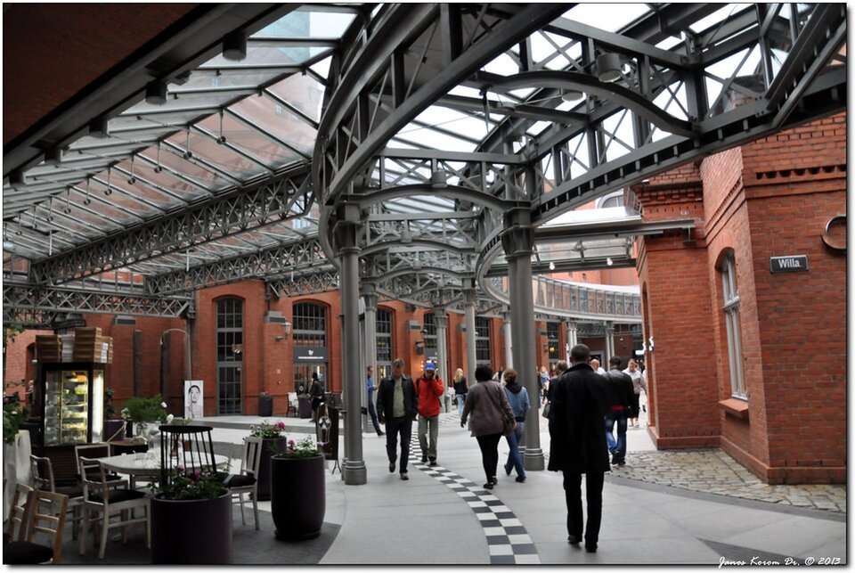 Zdjęcie przedstawia fragment budynku. Wykonany jest z czerwonej cegły. Nad przechadzającymi się tam ludźmi są stalowe, przeszklone konstrukcje. Po lewej stronie są wystawione stoliki i krzesła.  