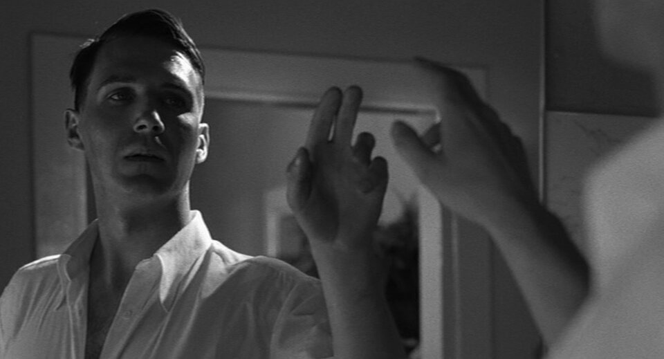 Zdjęcie przedstawia Gӧtha stojącego przed lustrem. Mężczyzna jest w rozpiętej koszuli. Ma uniesioną ku górze prawą dłoń.