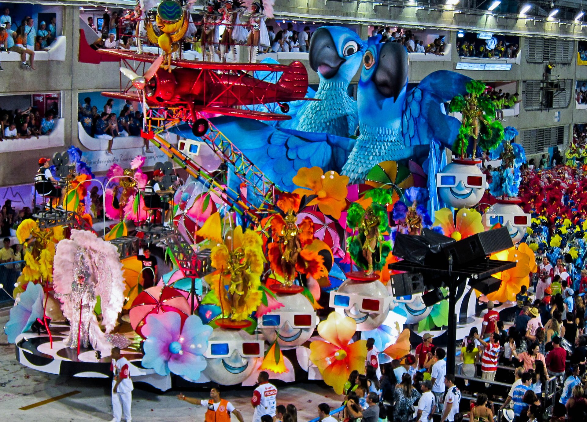 Zdjęcie przedstawia paradę, na której czele jest kolorowa platforma z dwoma wielkimi niebieskimi papugami
