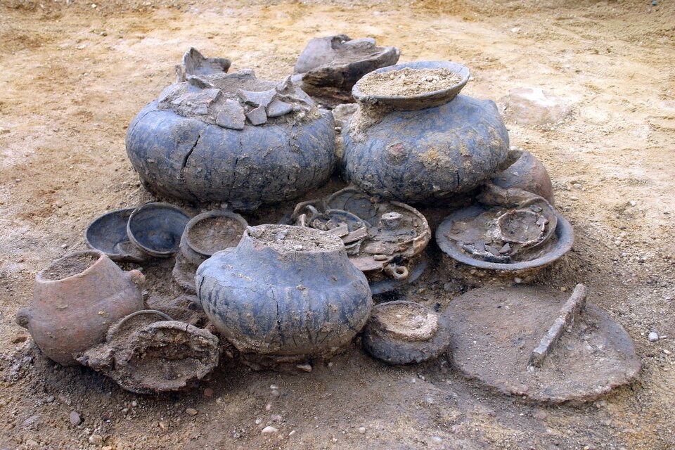 Zdjęcie przedstawia zbiór naczyń ceramicznych oraz metalowych przedmiotów odkrytych podczas wykopalisk archeologicznych, być może związanych grobem popielnicowym w kulturze halsztackiej. Naczynia są w różnym stanie zachowania – niektóre są uszkodzone lub częściowo zniszczone. Widać na nich ślady ziemi i osadów. Wśród obiektów znajdują się: duże pękate naczynia gliniane (z widocznymi pęknięciami i ubytkami), mniejsze miseczki i czarki, fragmenty metalowych przedmiotów, prawdopodobnie narzędzia lub ozdoby (np. fibule), płaski, okrągły metalowy obiekt z rączką – może to być pokrywka lub taca.